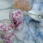 chopped red onions in mason jars ready to pickle with vinegar in measuring glass and slat nearby on marble counter