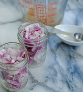 chopped red onions in mason jars ready to pickle with vinegar in measuring glass and slat nearby on marble counter