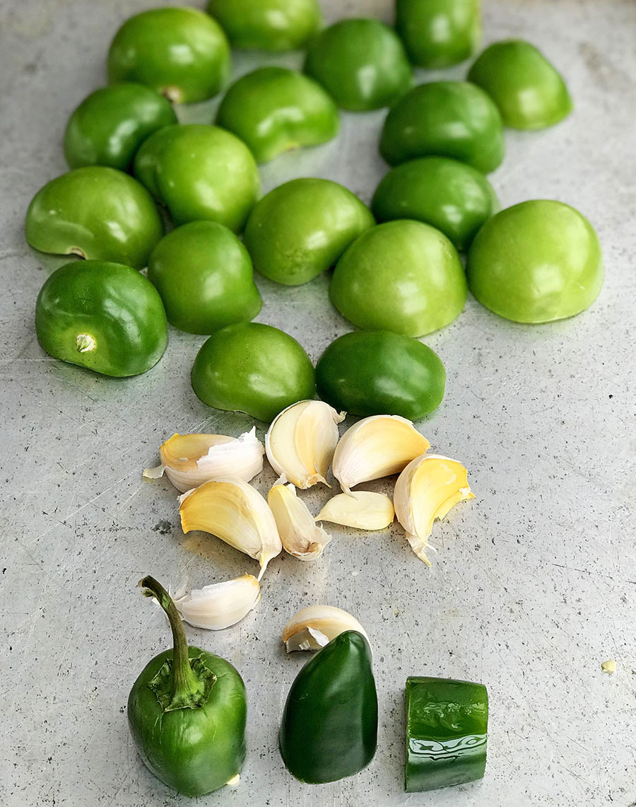 Metal sheetpan with jalapenos, garlic cloves, and halved tomatillos ready to roast. 