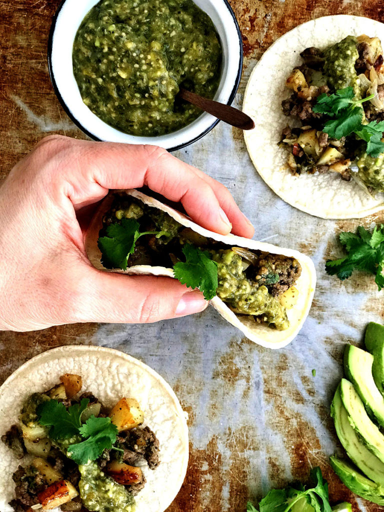 gluten-free taco in hand with avocado slices on a pan with corn tortillas