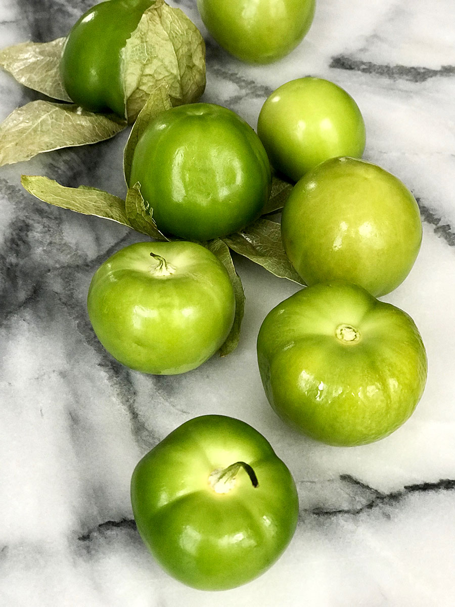 Eight bright green tomatillos wrapped and unwrapped strewn across marble counter. 
