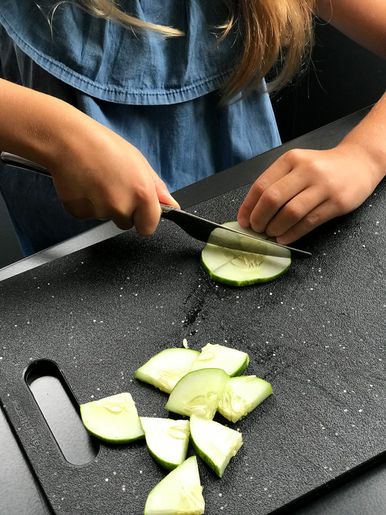 five year-old girl learning to cut a cucumber slice with a table knife on black cutting board