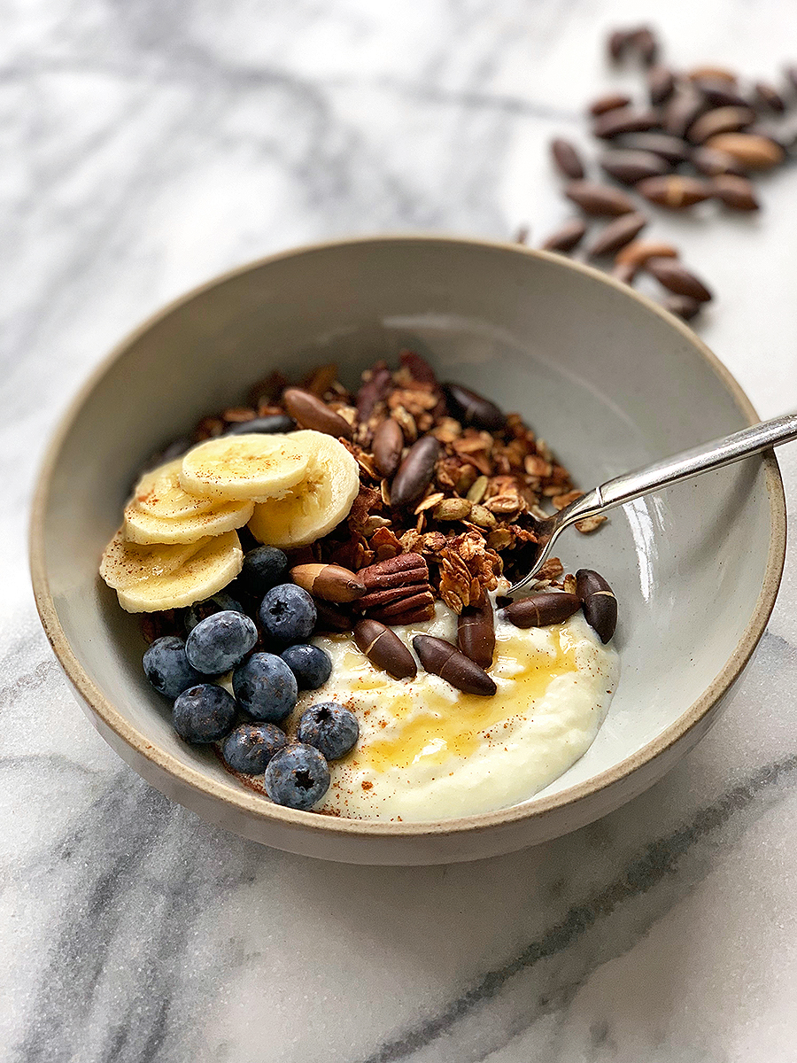 gray bowl with yogurt, honey, and crunchy granola on a marble table with toasted nuts scattered. 