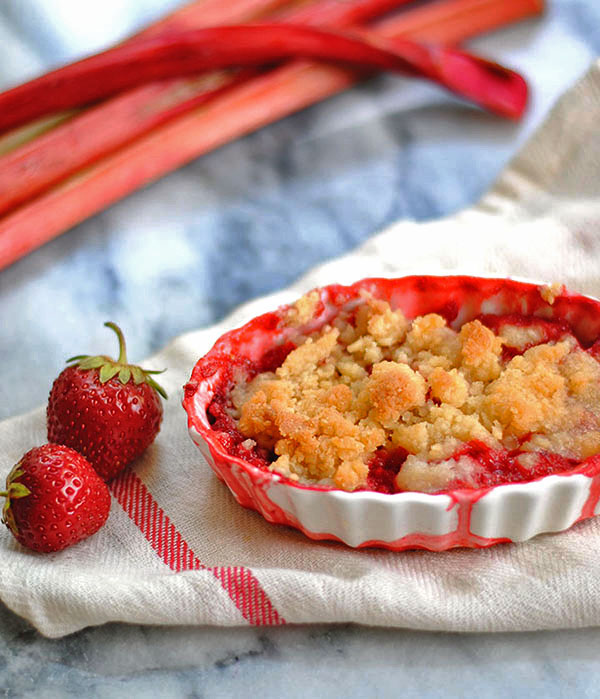 individual strawberry rhubarb almond crumbles in a white dish next to fresh rhubarb on a marble counter