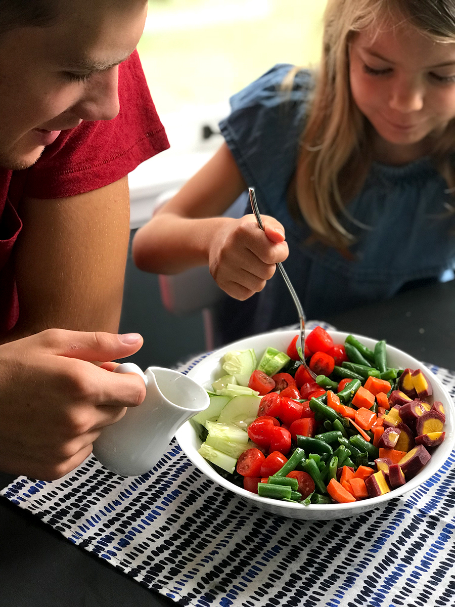 Teenage Cole with 5-year-old Evie digging into a gluten-free salad with dressing they made with fresh vegetables in a white bowl.