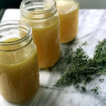 three mason jars with bone broth next to fresh herbs on marble slab