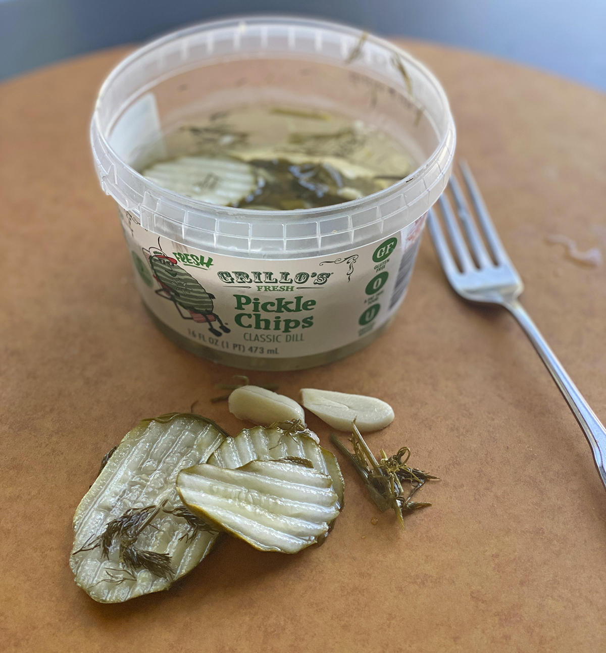 Grillo dill pickle container open with fork and pickle chips, dill, and garlic cloves on the counter. 