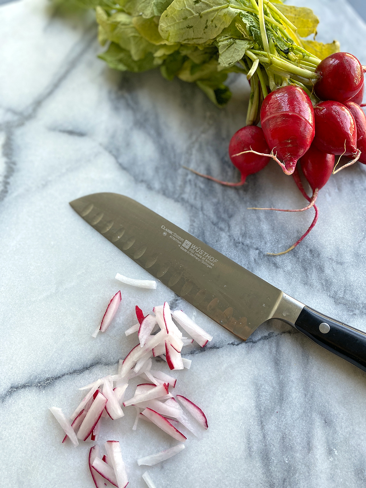 classic wusthof chef knife sitting next to chopped radish pile and a bunch of whole radishes on marble counter