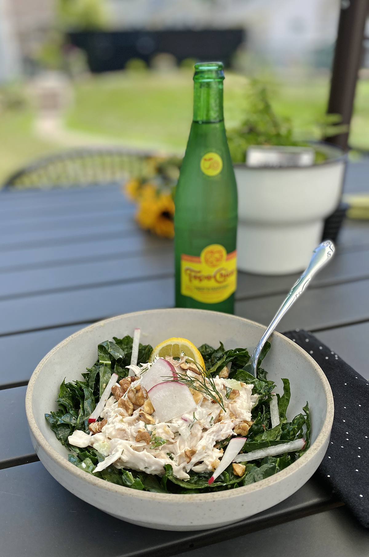 bowl of chicken salad in a bowl on a black table with black polka dot cloth napkin next to a cold bottle of Topo Chico sparkling water with plants in the background
