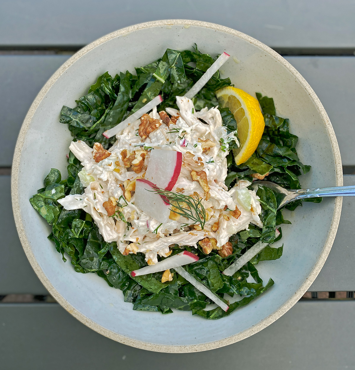 creamy chicken salad sprinkled with dill, radish, and walnuts on a mound of kale and radish slaw and a slice of lemon, all in a gray stone bowl on a black patio table. 