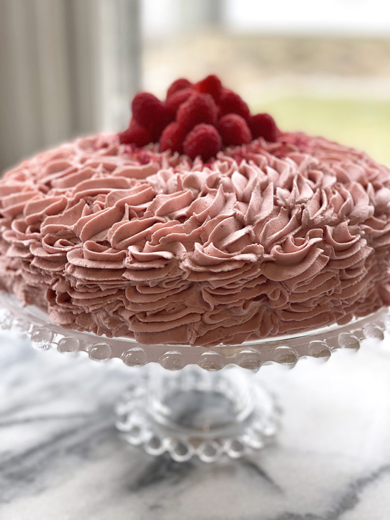 gluten-free raspberry frosted cake with fresh raspberries on top of a glass pedestal platter