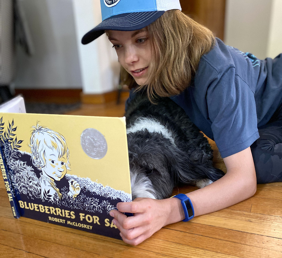 Eleven year old girl reading Blueberries for Sal children's book on the wood floor with her bernedoodle puppy looking on the pages and listening. 