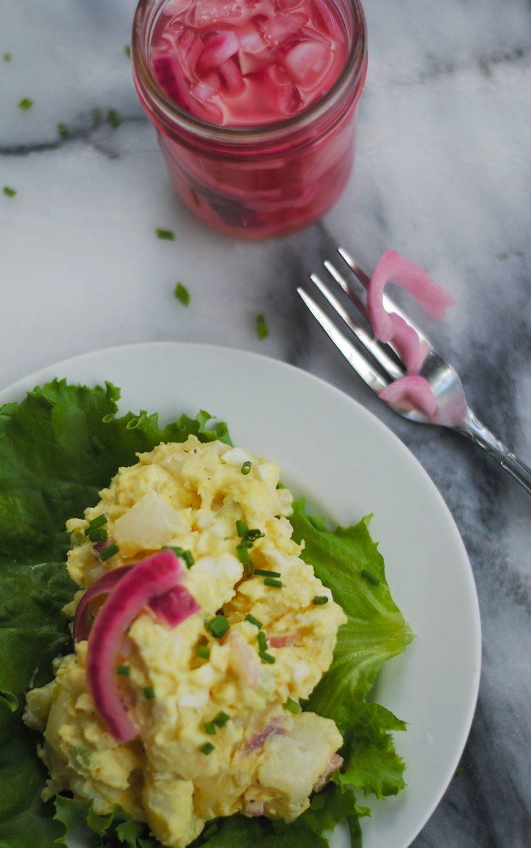 Classic yellow potato salad on green leaf of lettuce on white plate with pickled red onion strips on top with jar of pickled onions and fork on marble counter.