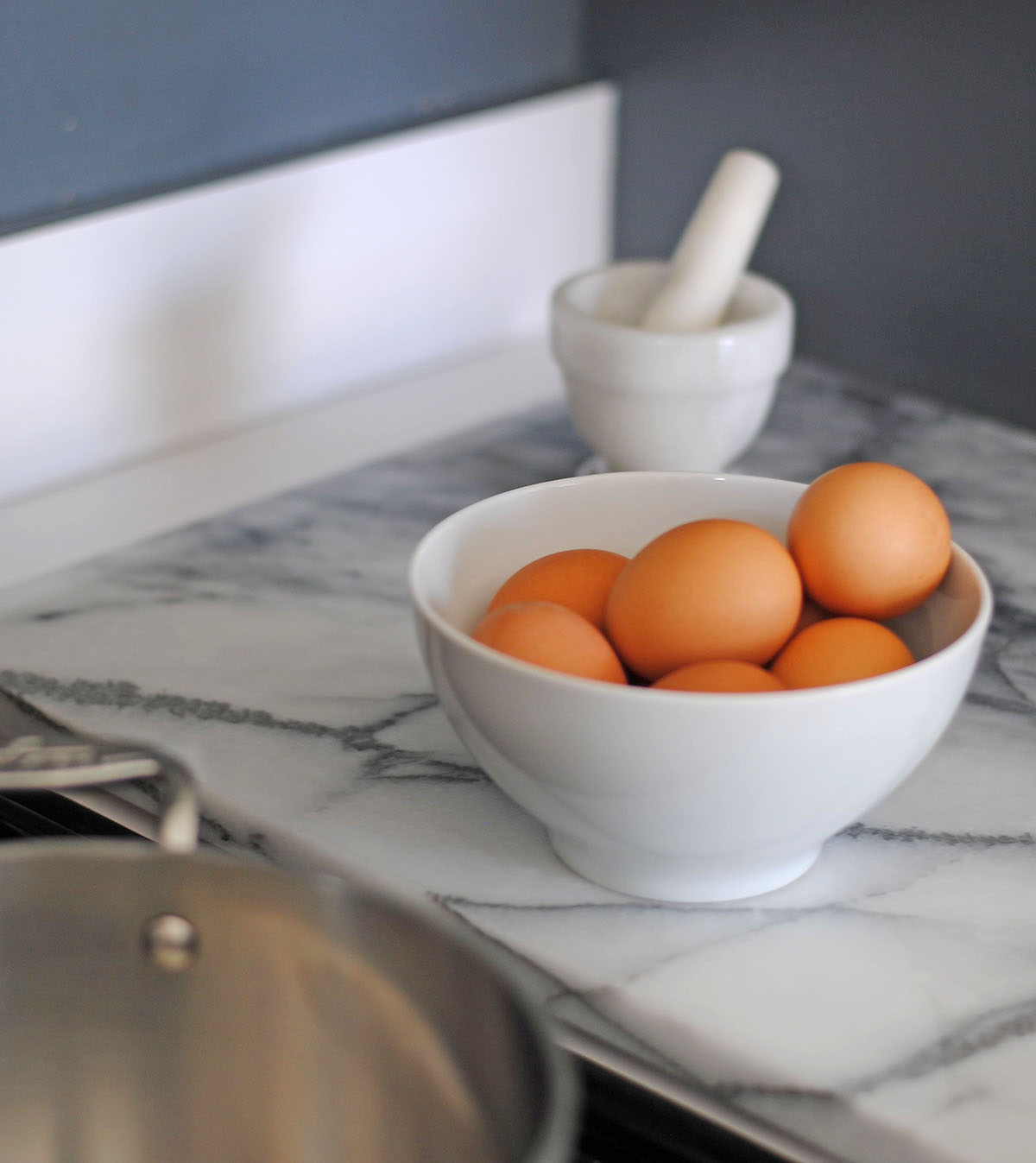 White bowl of fresh brown eggs on a marble counter ready to boil in a pot of water.