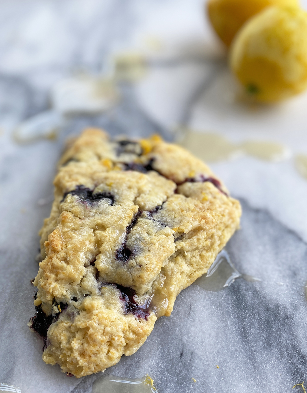 gluten-free blueberry scones with lemon glaze dripping off onto a marble surface and two zested lemons in the background