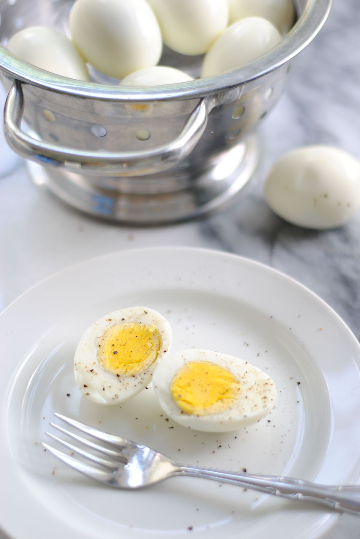 hard-boiled egg on white plate with salt and pepper next to colander with a bunch of peeled eggs