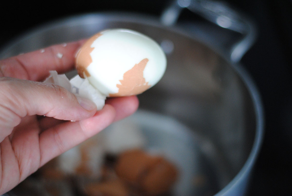 Hand peeling a smooth egg with the shell slipping off and a pot of shells in the background. 