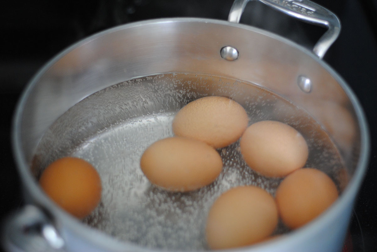 Metal pot of seven brown eggs in boiling water on stovetop. 