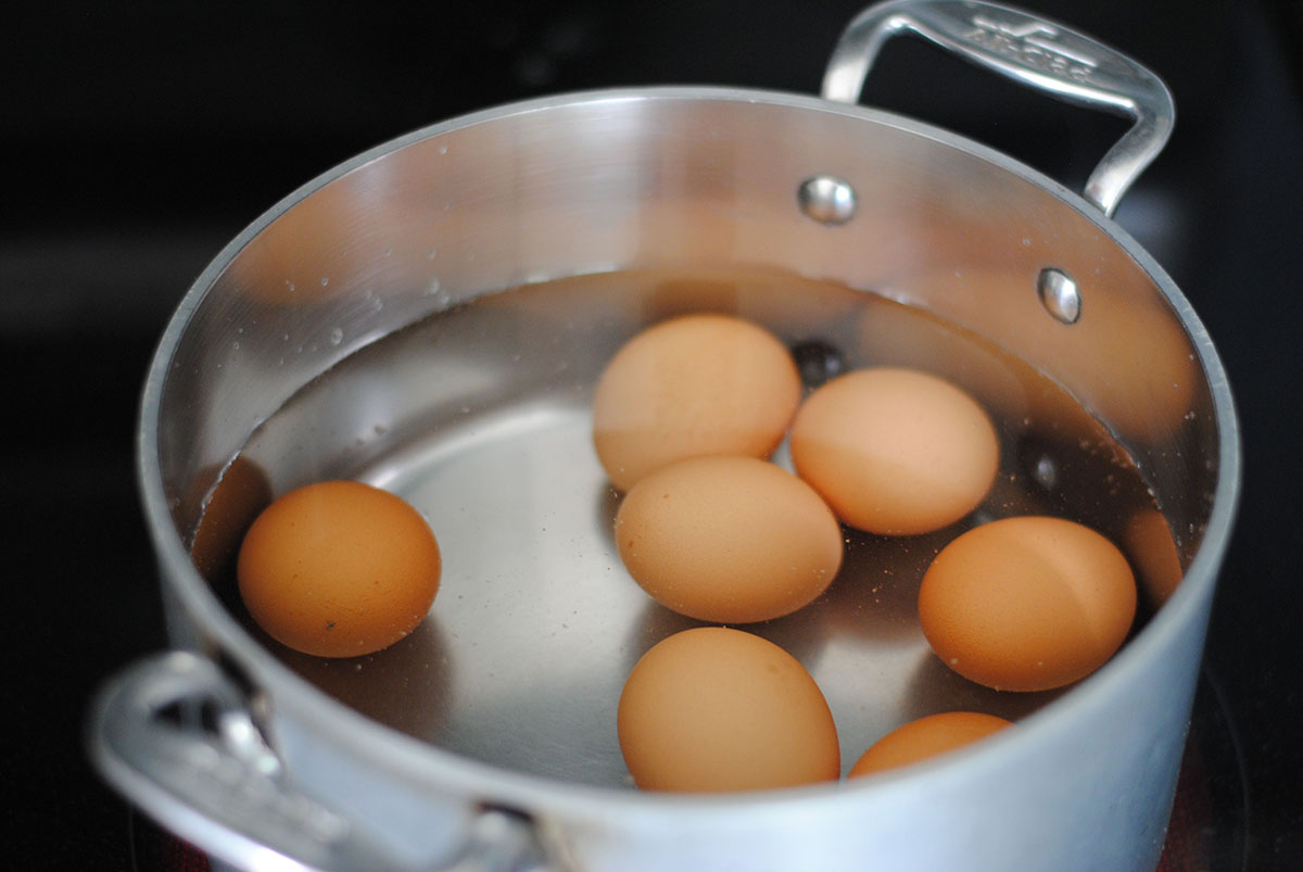 Silver pot of seven brown eggs covered in cold water ready to boil on the black stovetop. 