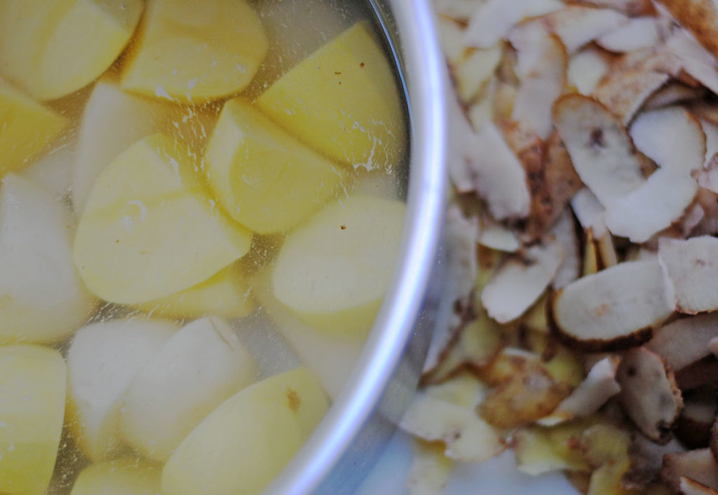 Pot of peeled potatoes in water ready to boil for potato salad next to pile of potato peels on marble counter. 
