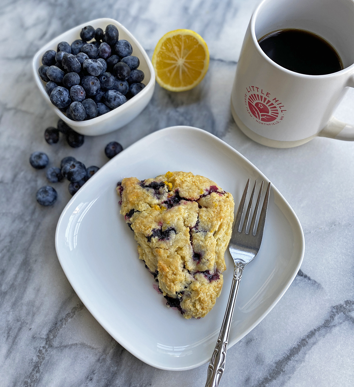 white plate with a blueberry scone with fork, cup of coffee, dish of blueberries and half of a lemon on a marble table