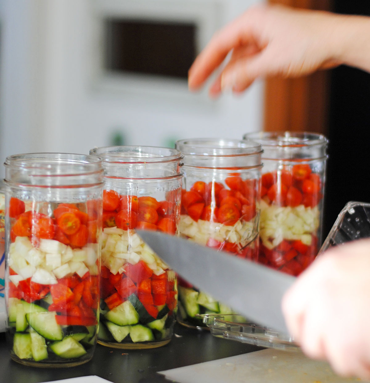 four mason jars lined up with Greek salad ingredients from cucumber red pepper, fennel, and cherry tomatoes and a knife held by a hand prepping. 