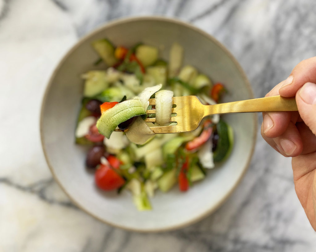 A gold fork taking a bite of raw zucchini, kalamata olives, red peppers, and tomatoes above a bowl of freshly chopped ingredients on a marble counter. 
