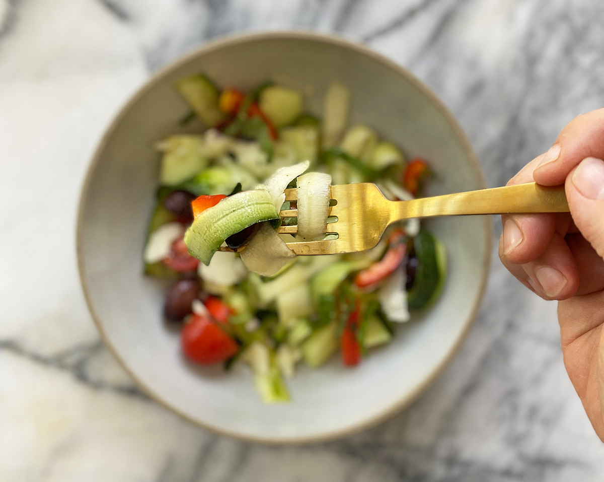 A gold fork taking a bite of raw zucchini, kalamata olives, red peppers, and tomatoes above a bowl of freshly chopped ingredients on a marble counter.