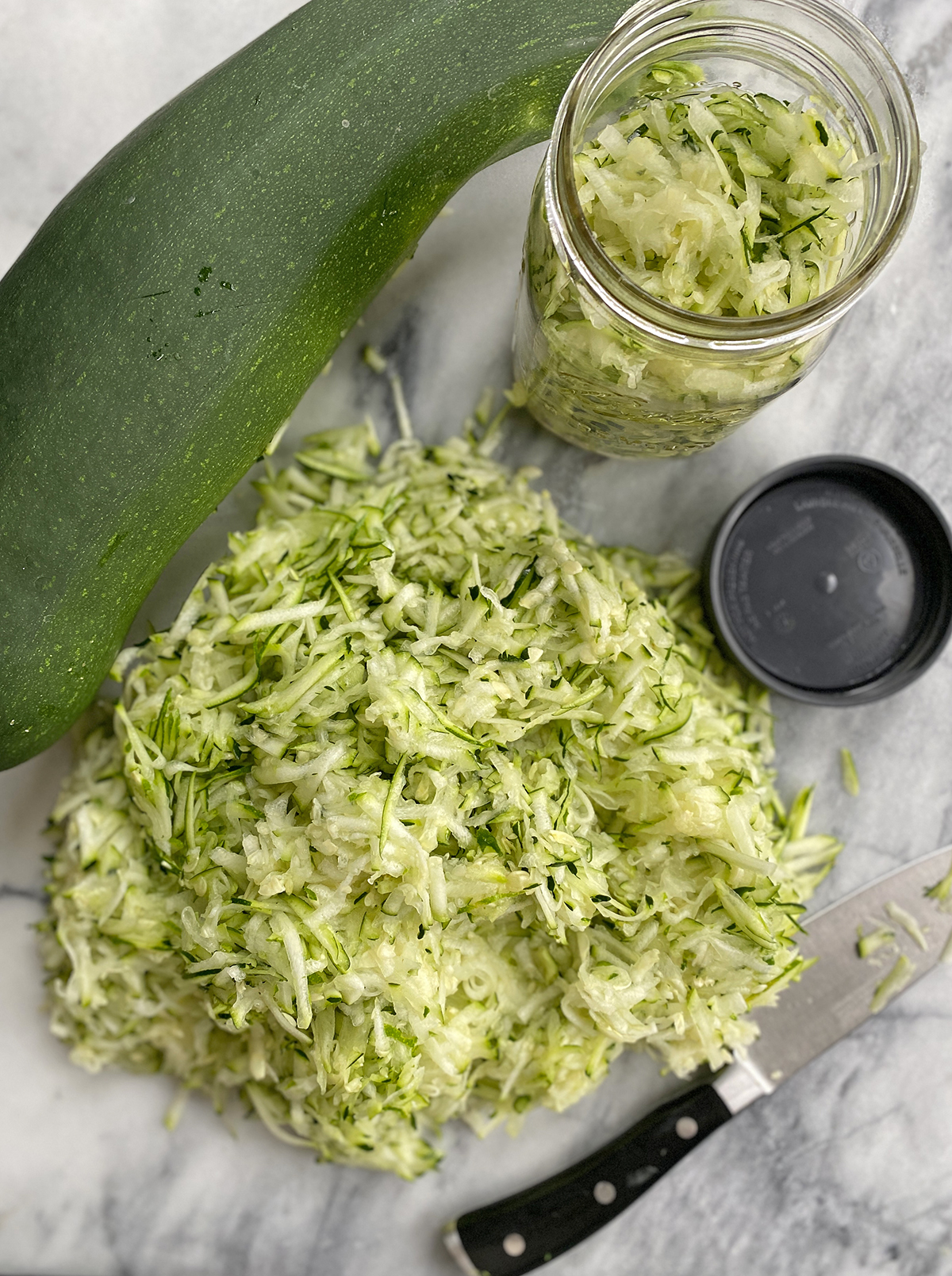 A pile of shredded zucchini sitting next to freezer jar with lid and a large green zucchini on a marble counter. 