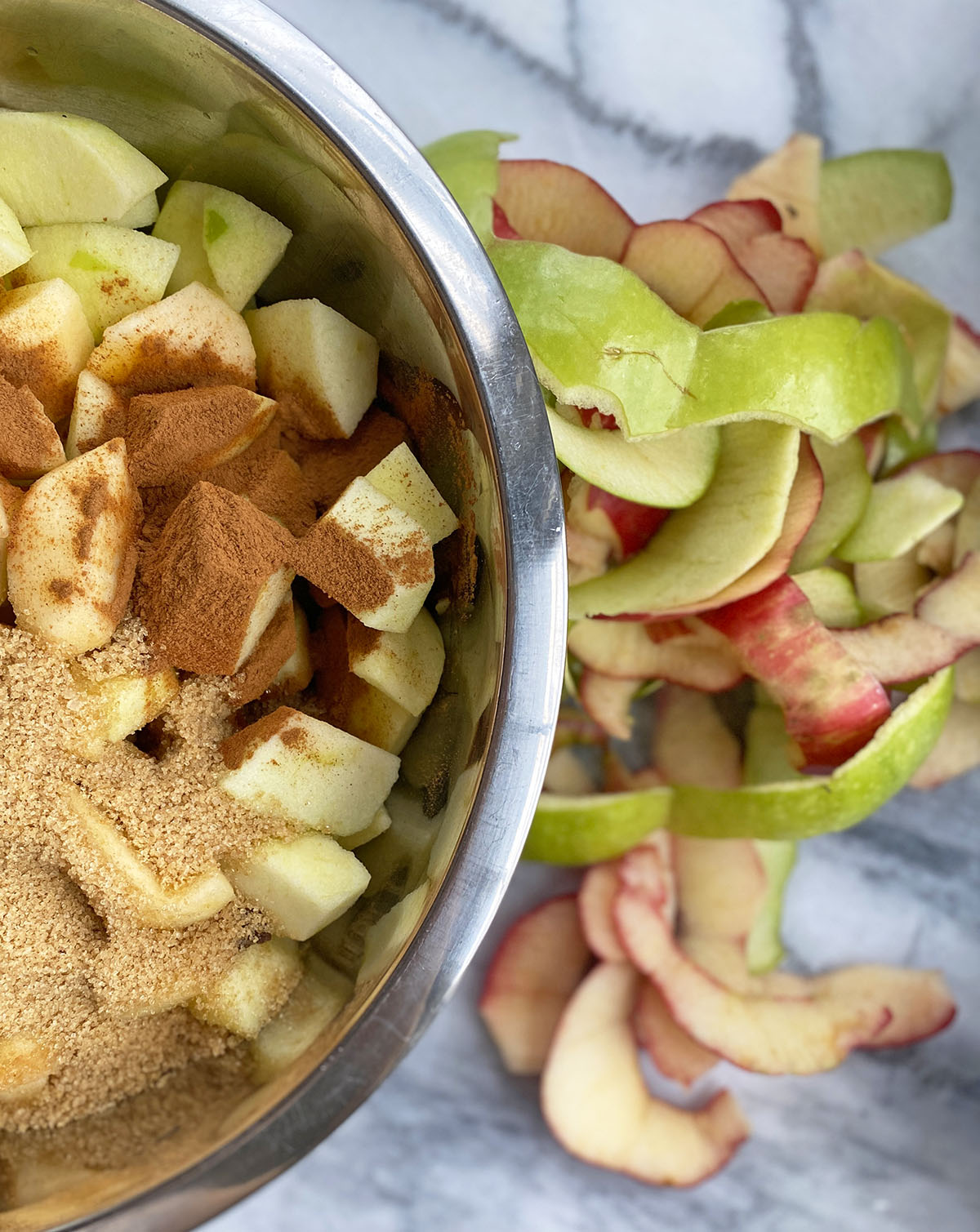 Large silver mixing bowl with apple chunks, cinnamon, and brown sugar for making apple crumble next to pile of peels on a marble counter. 