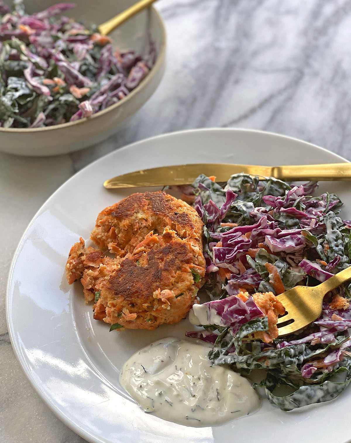 White plate with gluten-free salmon cake next to red cabbage and kale slaw with gold fork and bowl full of extra slaw on marble table. 