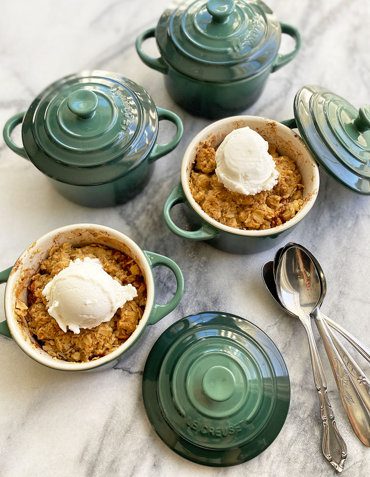 Individual mini le creuset green baking dishes with apple crumble and vanilla ice-cream and spoons sitting on marble counter. 