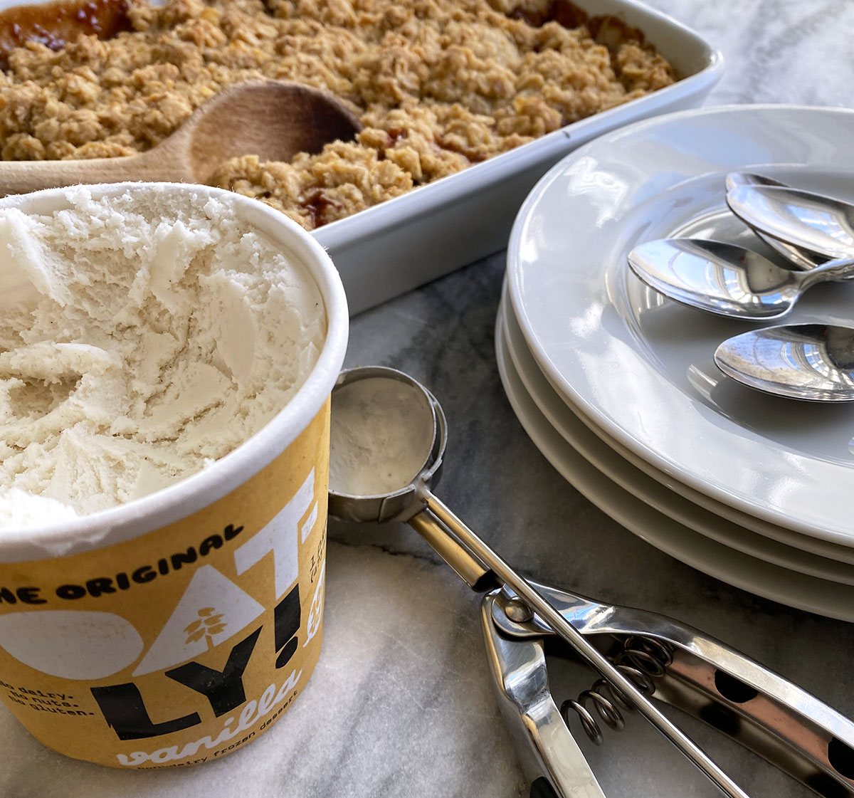 Open tub of vanilla Oat-ly ice cream, scoop, plates, and baking dish of apple crumble on marble counter. 