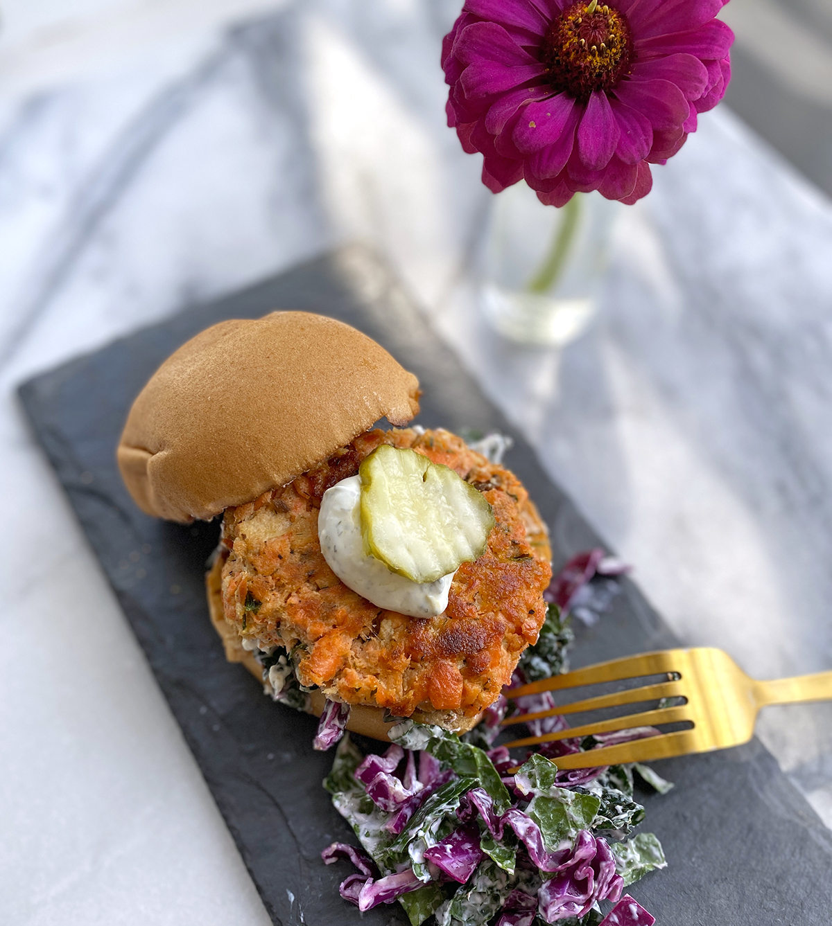 Gluten-free salmon cake in a bun with yogurt dip, pickle, red cabbage slaw, on a black plate and magenta colored Zinnia in a small glass vase. 
