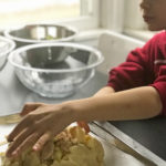 Child's hands patting together an apple tart on a baking sheet next to glass bowl.