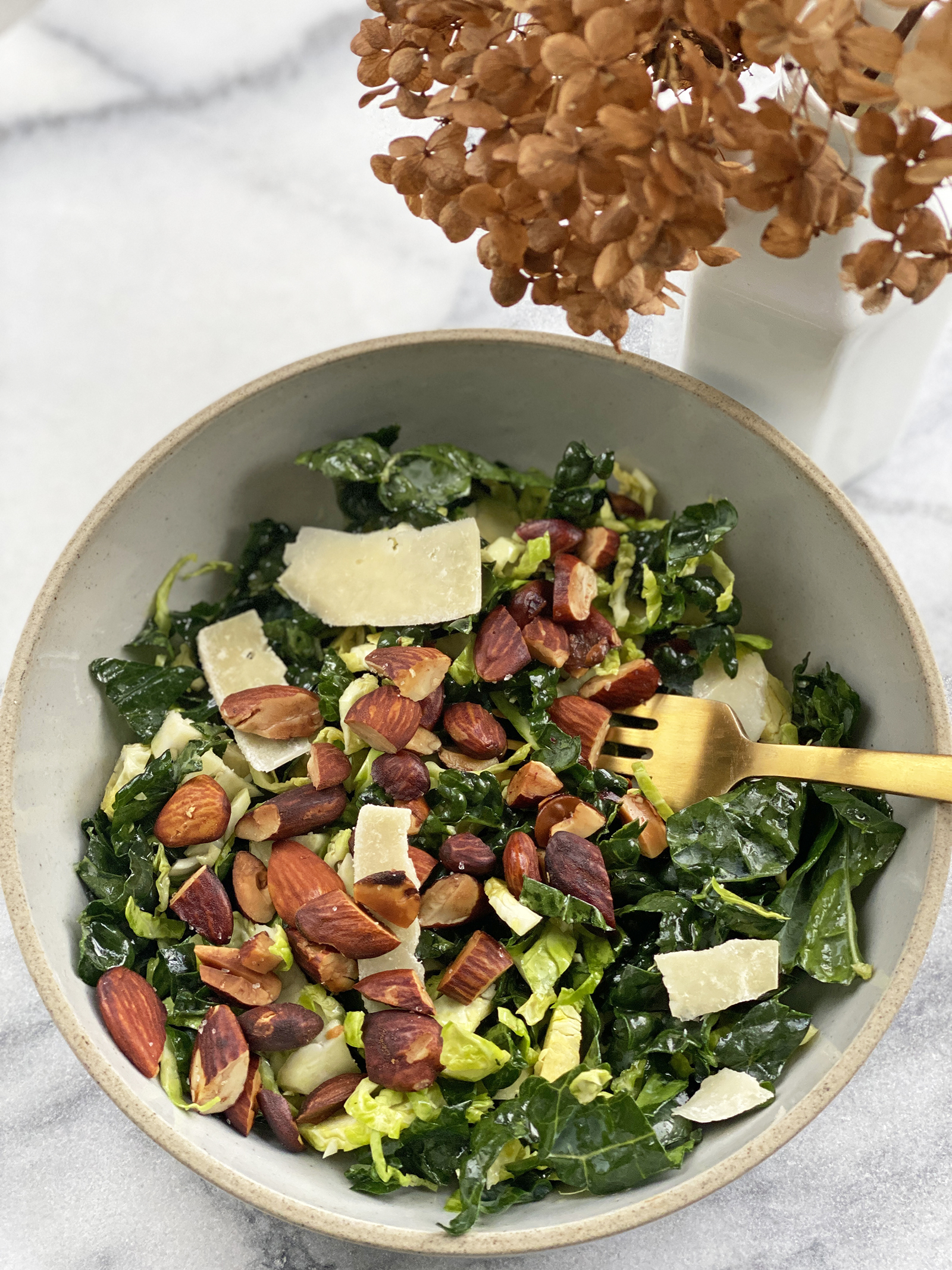 stone bowl of kale and Brussels salad with pecorino shavings, almonds, with gold fork on marble table. 