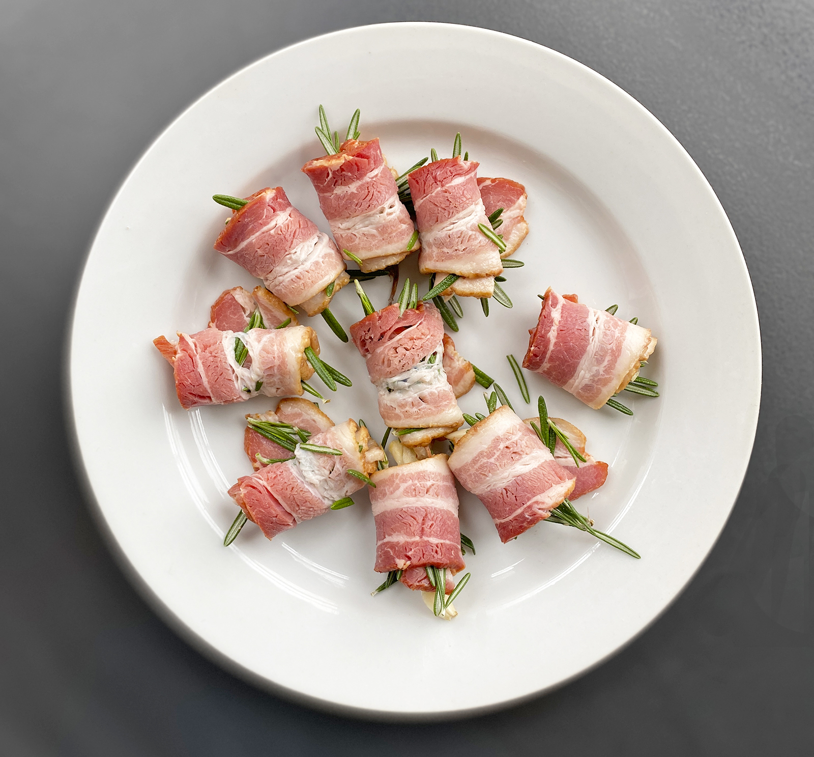 white plate of rosemary and garlic cloves rolled into bacon pieces on the counter. 