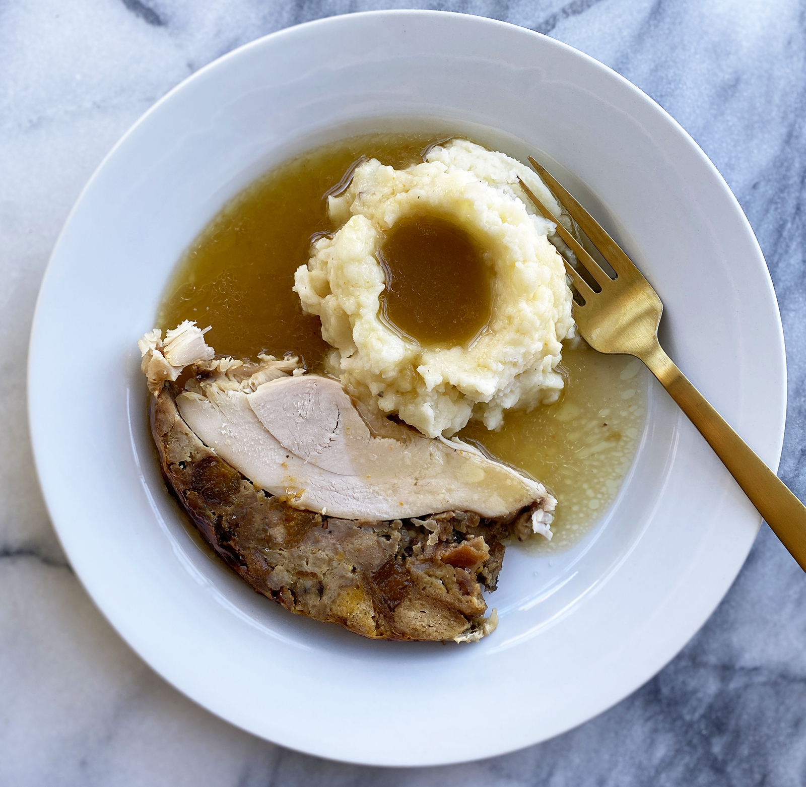 white plate on marble surface with gold fork sticking into mashed potatoes next to turkey and stuffing with gravy