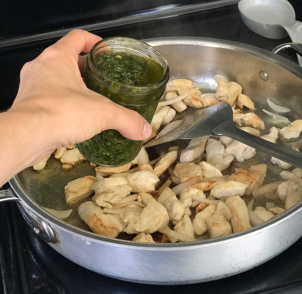 chicken cooking with hand holding mason jar of green sauce ready to pour in pan