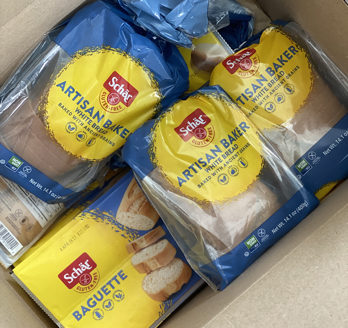 Cardboard box full of Schar gluten-free bread and baguettes on top of other gluten-free groceries. 