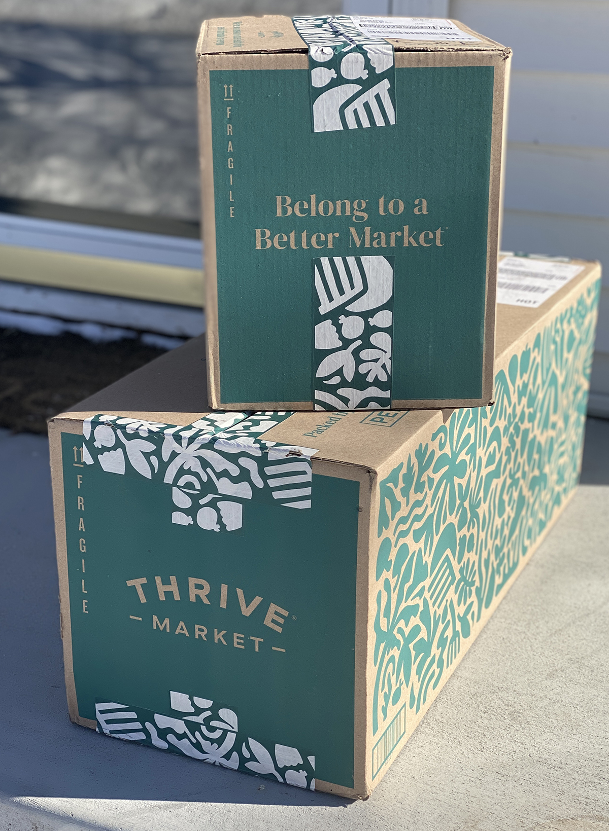 Two green Thrive Market boxes stacked on front step with sun shining on them in front of the door with gold trim. 