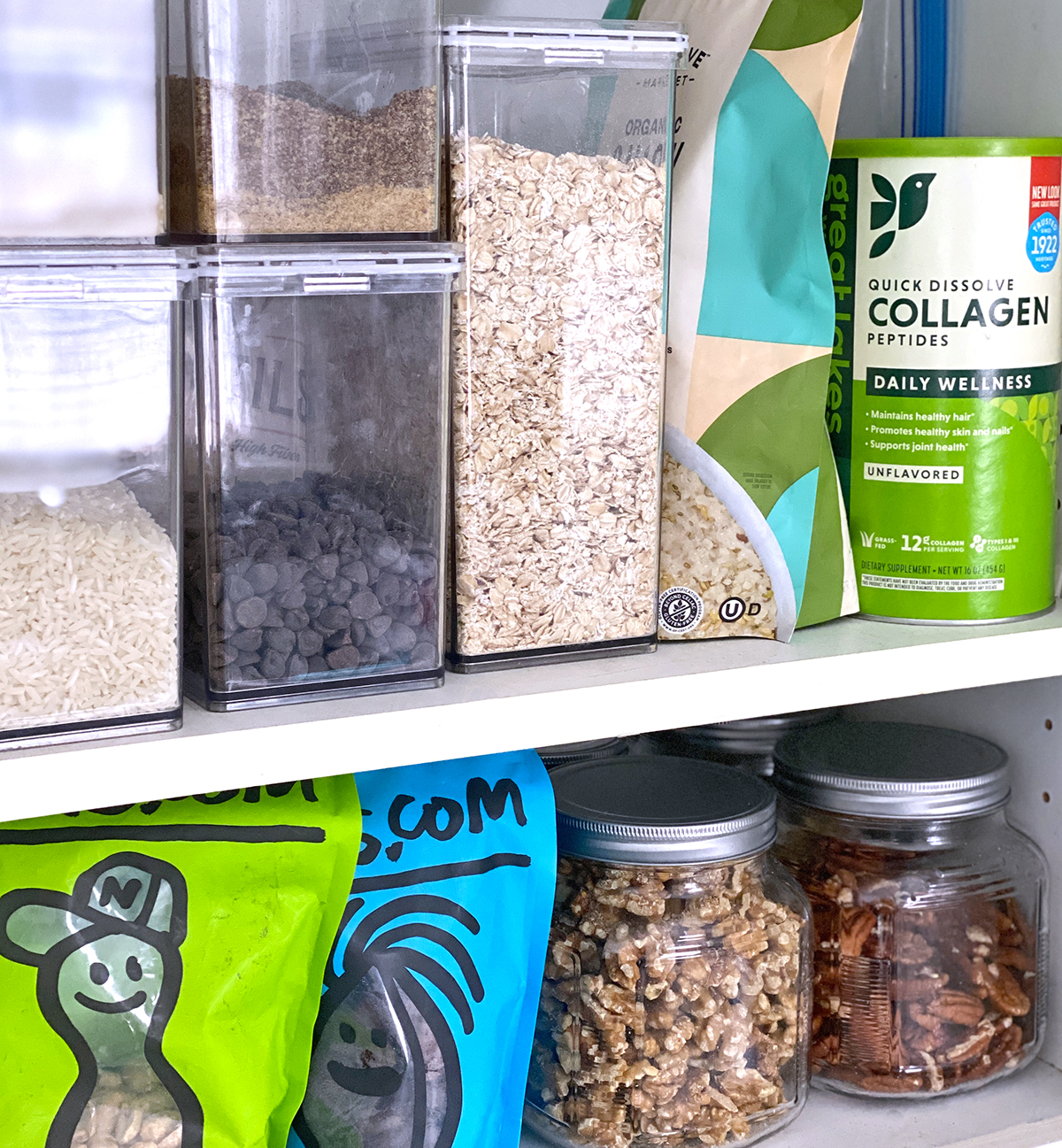 White cupboard opened to clear containers of rice, chocolate chips, oats, flaxseed, and quinoa next to collagen peptides and a shelf of jars full of pecans, walnuts, and bags of peanuts and apricots. 