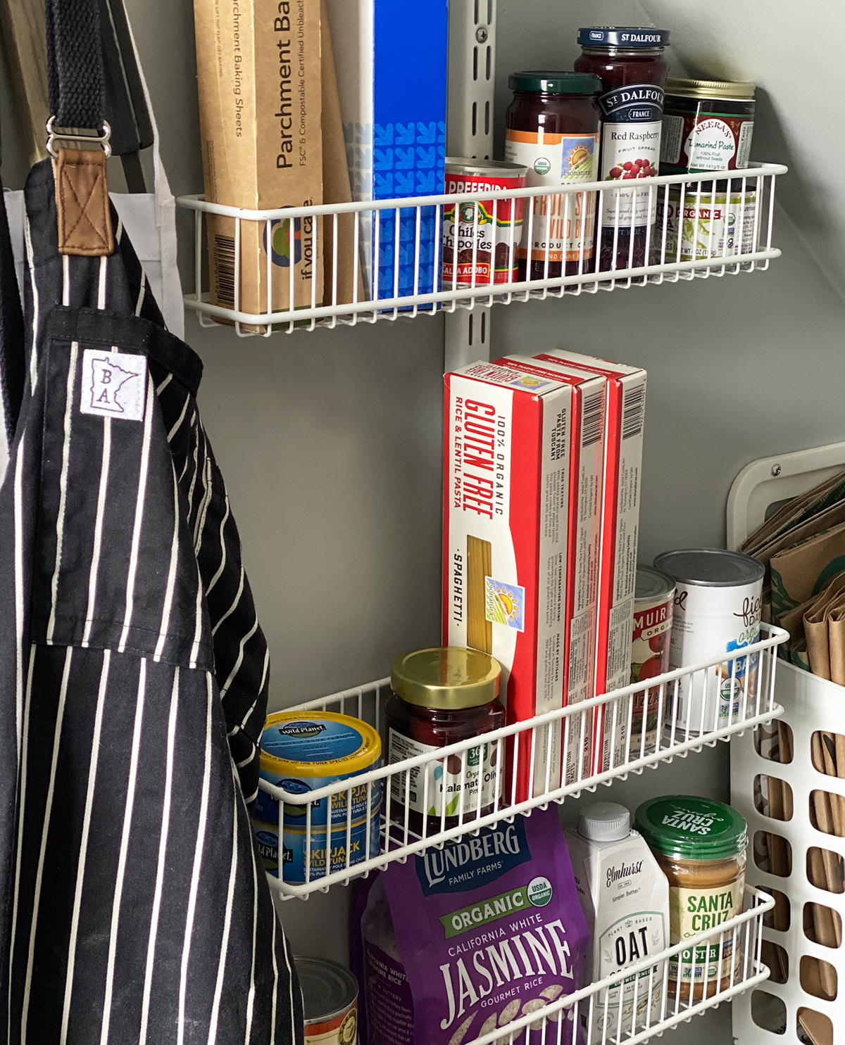 Aprons hanging next to white wire pantry shelves on gray wall with supplies and gluten-free groceries like jam, pasta, rice, and peanut butter. 