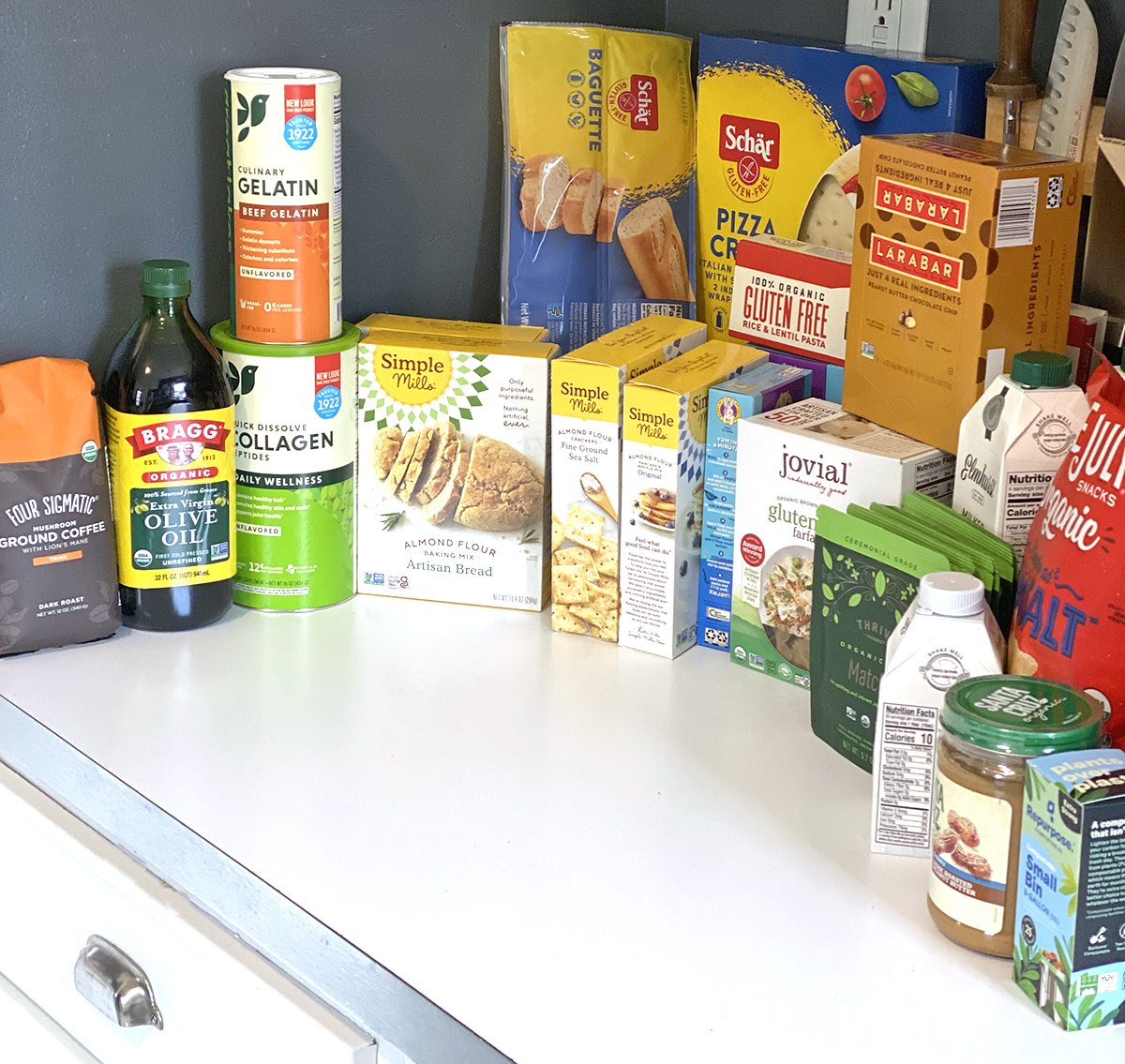 White counter with boxes of gluten-free food from Thrive Market started with coffee, olive oil, collagen, bread, crackers to matcha, peanut butter, and chips. 