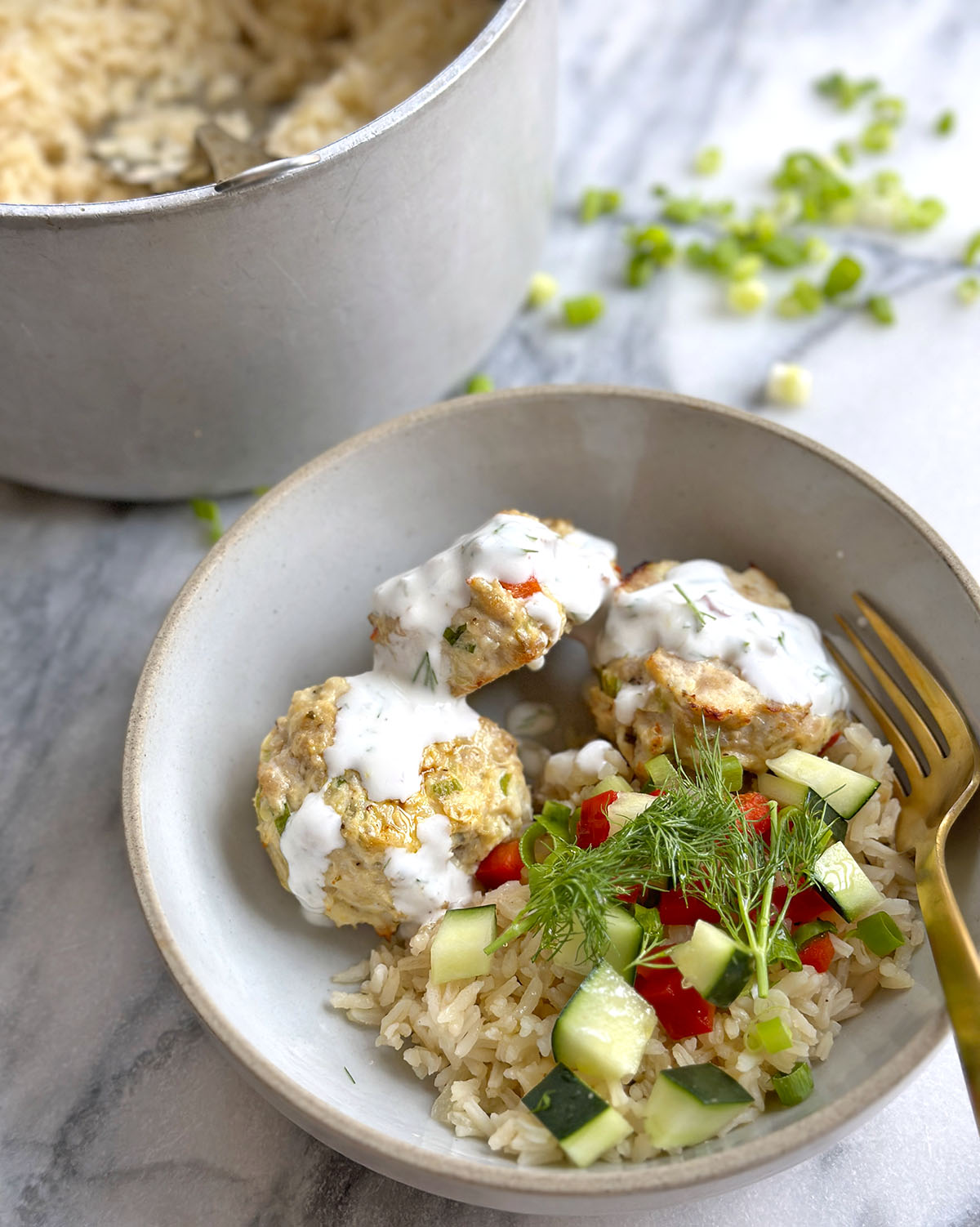 A bowl of rice, three meatballs, yogurt sauce, and chopped red pepper and dill salad on top. 
