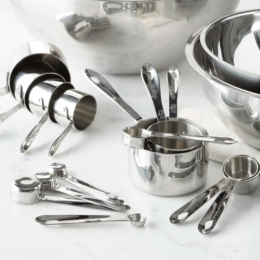 seven measuring cups displayed next to teaspoons and tablespoons in front of a silver set of empty bowls on a marble counter