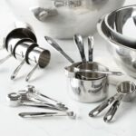 seven measuring cups displayed next to teaspoons and tablespoons in front of a silver set of empty bowls on a marble counter