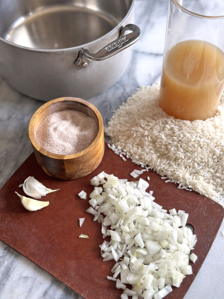 Silver pot, chicken broth in a glass pitcher, white rice, white onion on a cutting board, two garlic cloves and salt sitting on a marble counter.
