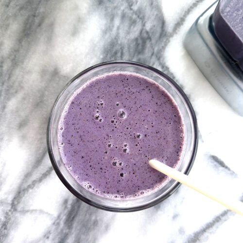 Blueberry smoothie with straw sitting next to blender on a marble counter.