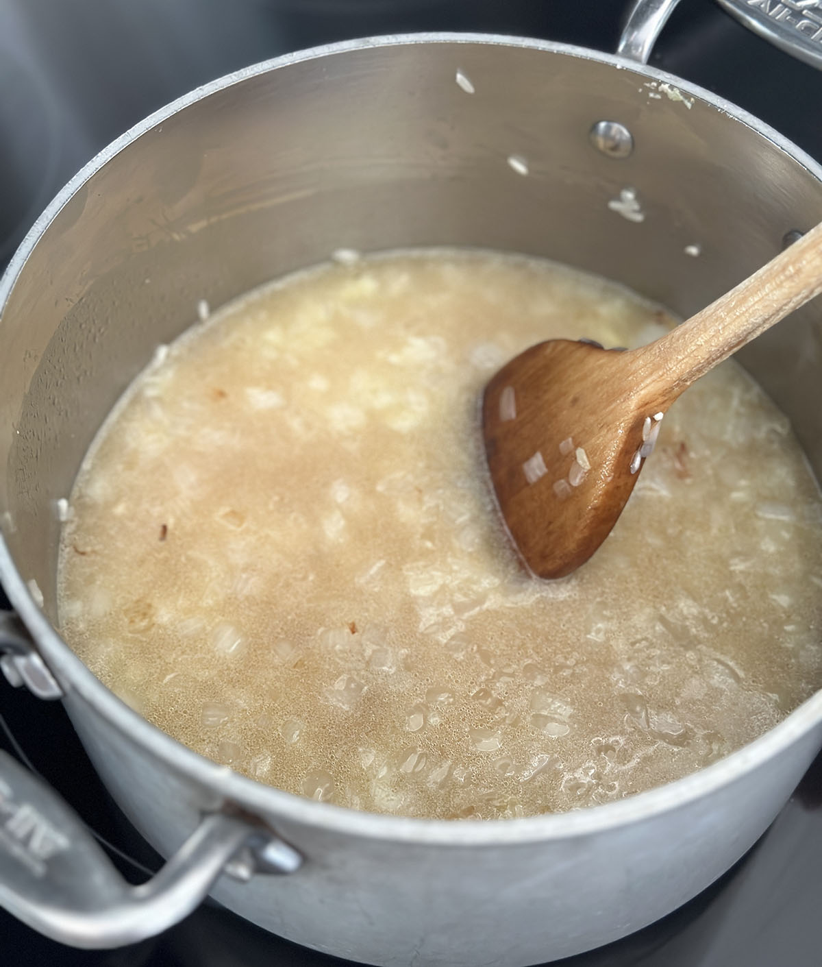 Wood spoon stirring broth into rice and onions in a silver pot. 